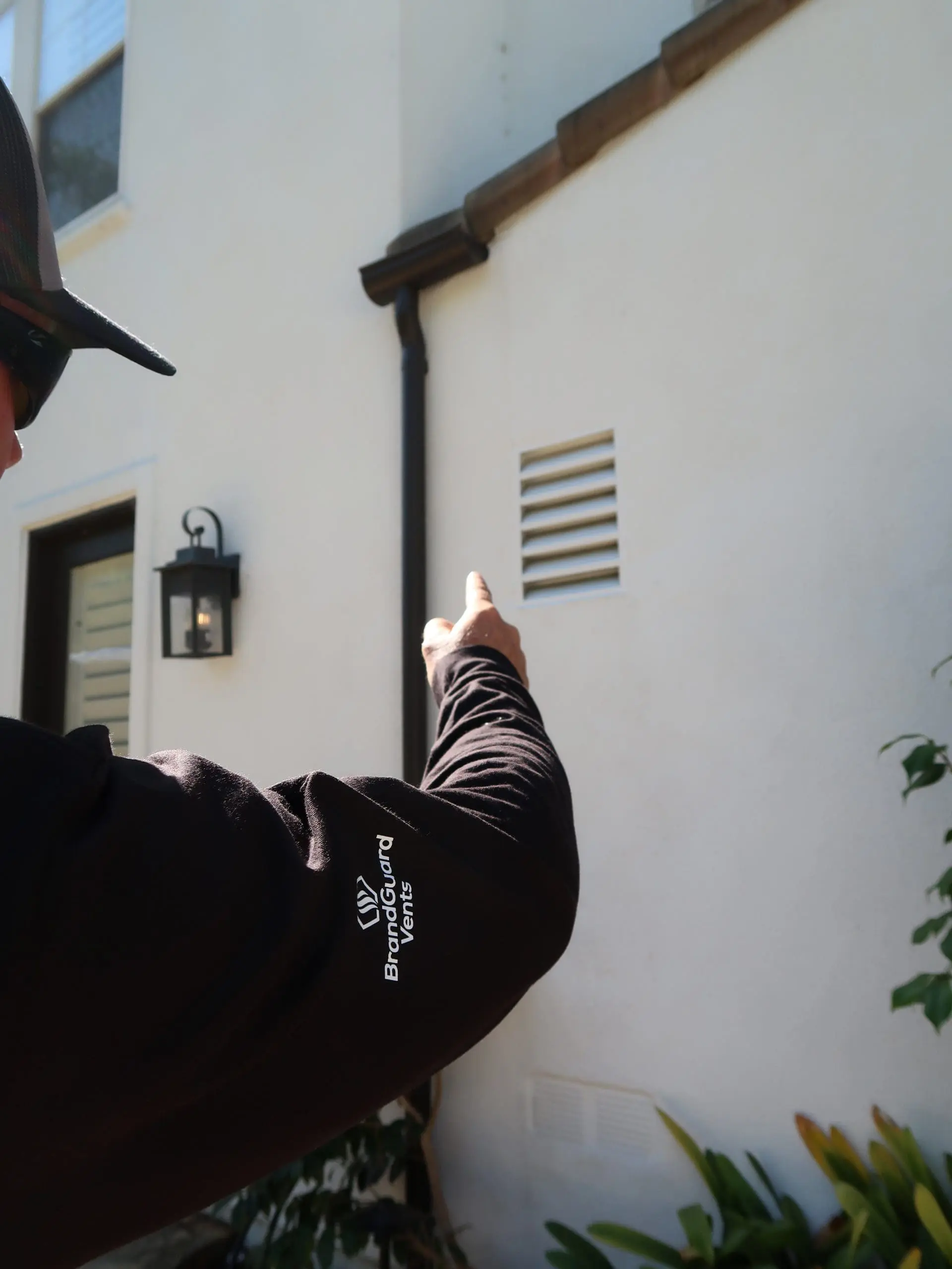 Person in a black shirt pointing at a vent on a white exterior wall near plants and a black lamp.