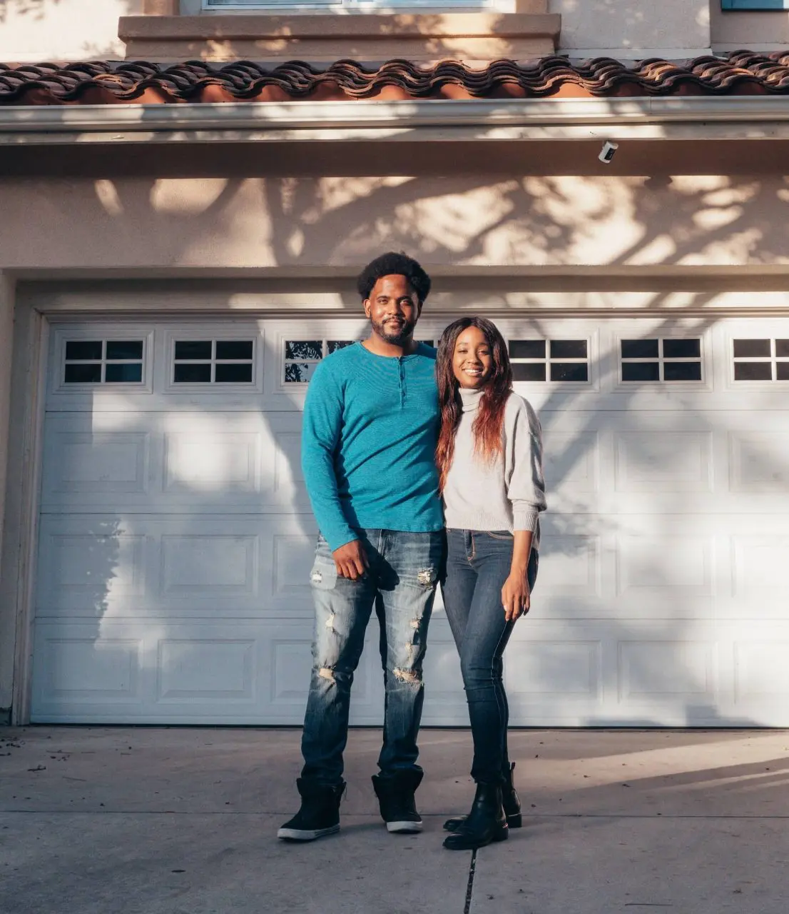Two people smiling in front of a white garage door on a sunny day.