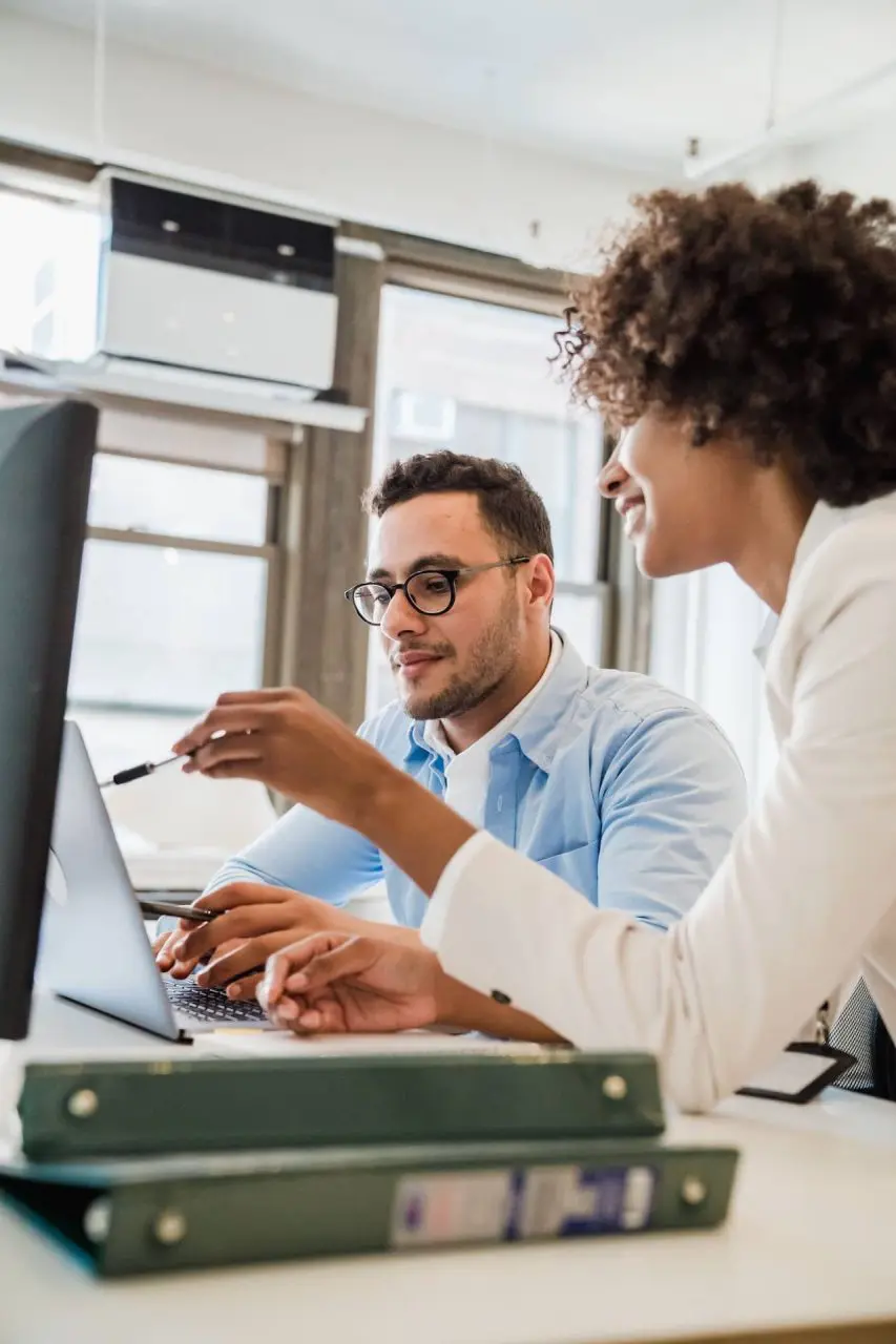 Two colleagues work together at a desk, viewing a computer screen in an office.