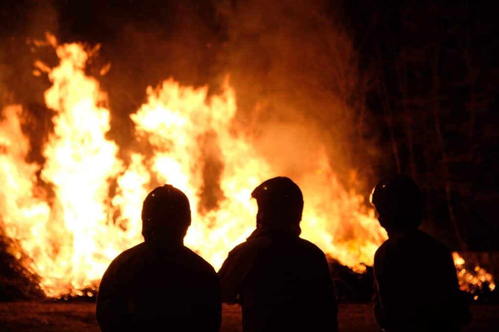 Silhouettes of three firefighters standing in front of a blazing wildfire at night.