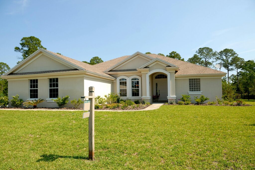 Single-story white stucco house in California with a "for sale" sign in the front yard. Trees and blue sky in background.