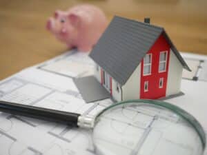 Model house, piggy bank, and magnifying glass next to house blue print papers.